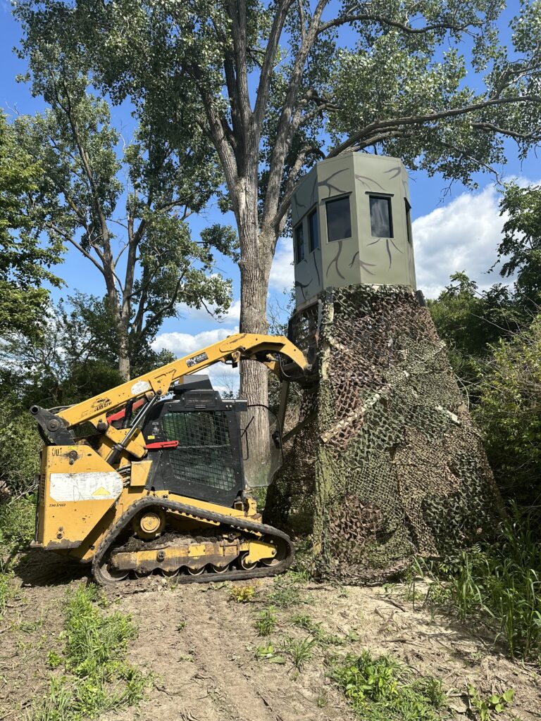 Tracked loader lifting a camouflaged hunting blind in a wooded area